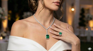 A woman wearing an off‑shoulder white dress showcases a diamond necklace with a large emerald pendant and a matching emerald ring.