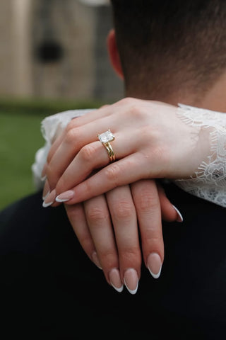 Close up of a bride and groom holding hands, featuring a radiant cut diamond engagement ring paired with a wide yellow gold wedding band against intricate white lace.