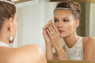 Woman wearing diamond headband, statement diamond ring and drop earrings while looking into a mirror in soft bridal lighting