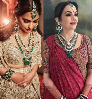 Isha & Nita Ambani traditional Indian attire, both styled in emerald and diamond jewelry featuring layered necklaces, earrings, and maang tikka; one in a silver lehenga and the other in a deep red saree.