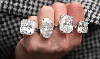 A hand wearing four large diamond rings in different shapes — cushion, oval, and square cuts — showcasing oversized stones set in simple prong settings against a checkered fabric background.