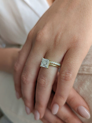 Close up of a hand wearing a princess cut diamond solitaire ring in yellow gold, paired with a delicate pavé diamond wedding band against a soft neutral background.