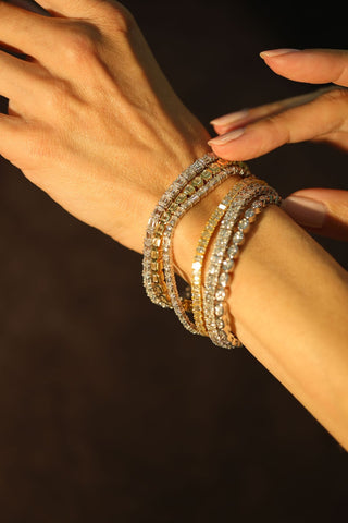 Close up of a woman’s wrist layered with diamond tennis bracelets and mixed cut diamond bangles, illuminated by warm golden light.